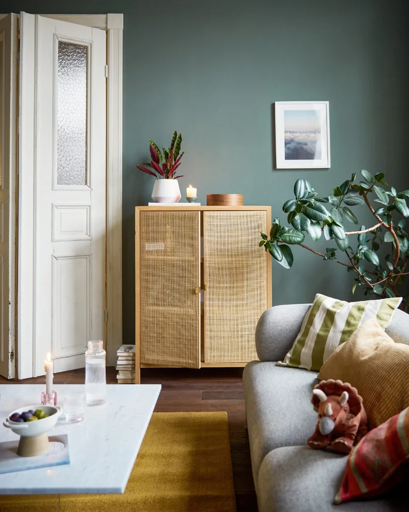 Interior shot of a living room with green walls, a light wood rattan cabinet, a grey sofa with pillows, and a white coffee table.