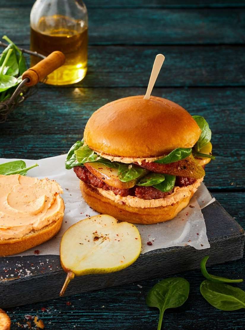 Gourmet burger featuring beef, pear slices, greens, and sauce, presented on a rustic wooden board with spinach and olive oil.