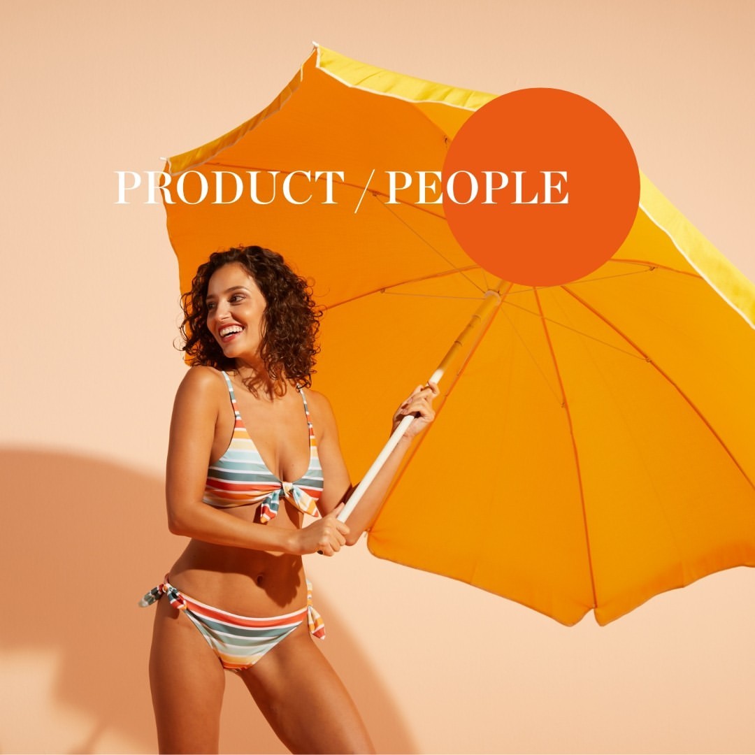A woman in a striped bikini stands smiling in front of a peach background while holding an open yellow umbrella.