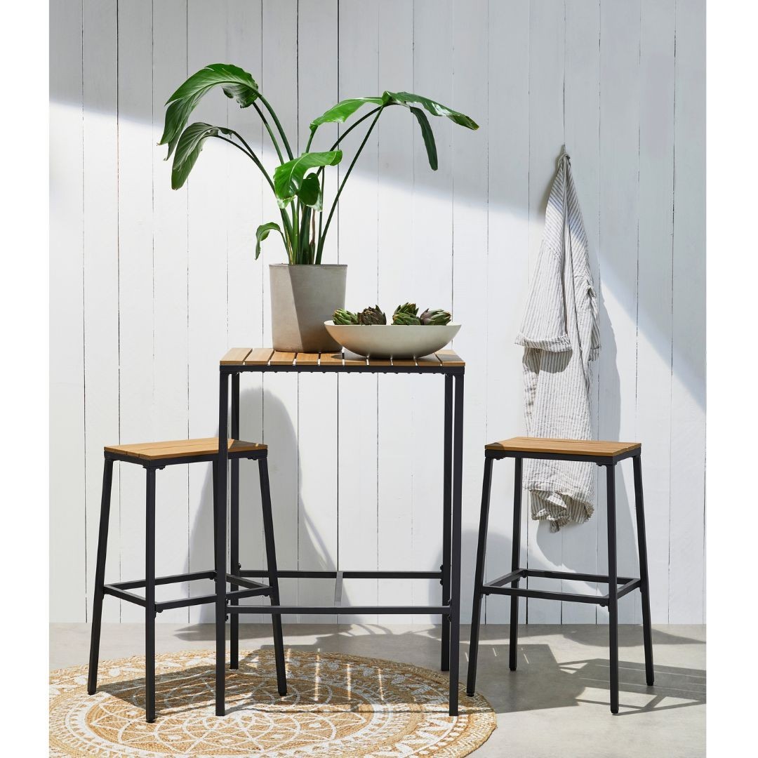 Outdoor setting with a small wooden table, two stools, potted plant, bowl with artichokes, and a white wooden panel background.