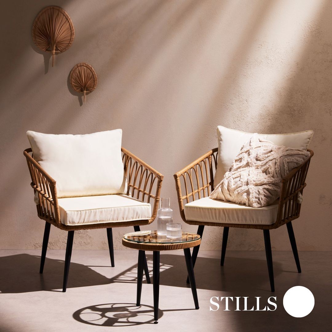 Two wicker chairs with cushions and a small table are bathed in warm light, set against a neutral wall adorned with woven fans.