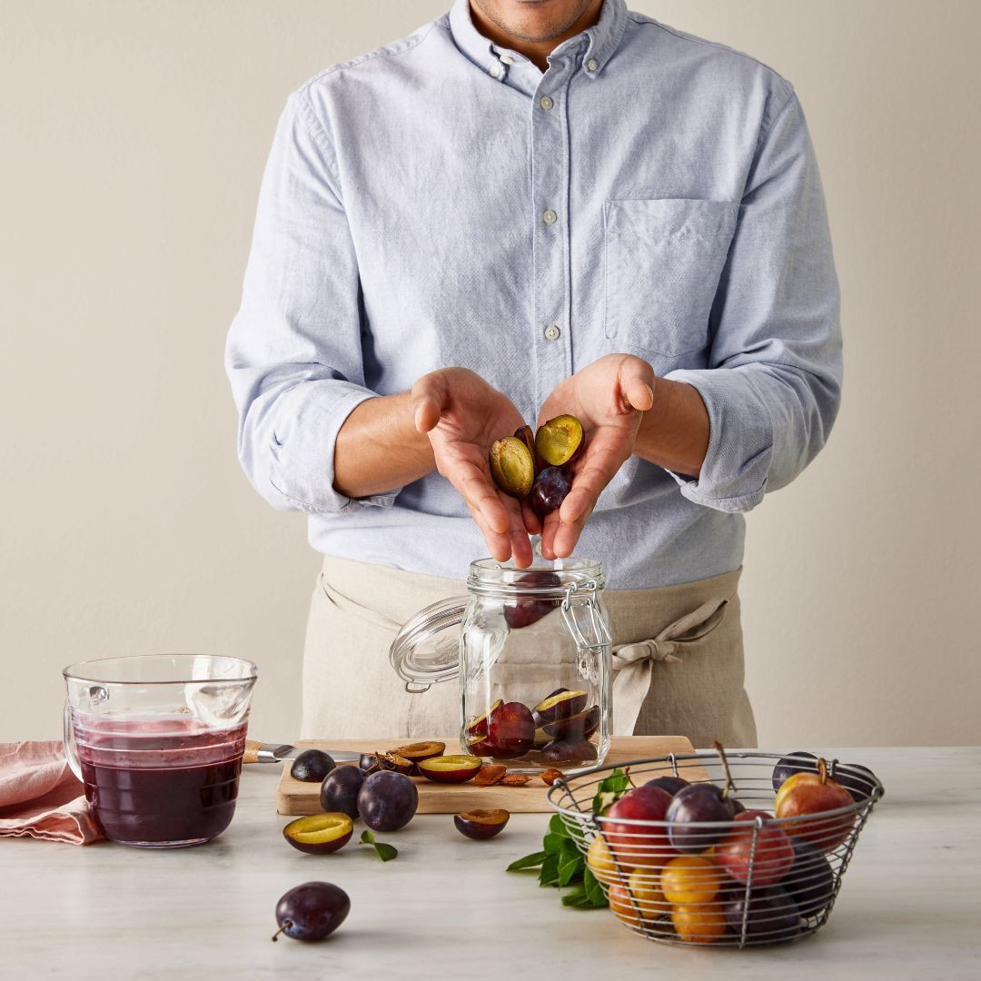 Man filling a glass jar with fresh plums on a marble countertop. A cup of plum juice, wire basket with plums, and cutting board are visible.