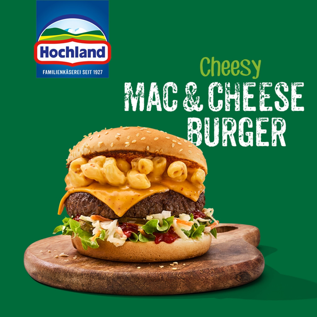 A close-up of a 'Mac & Cheese' burger, stacked high on a wooden board against a green background, showcasing layers of beef, cheese, and macaroni.