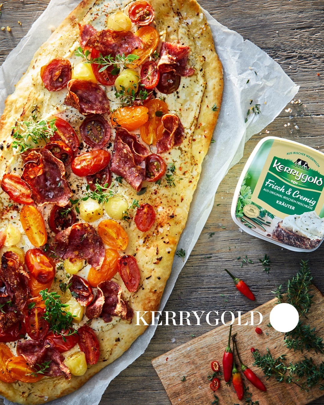 Overhead shot of flatbread pizza with tomatoes, salami, and herbs on parchment paper with a container of Kerrygold cheese nearby.