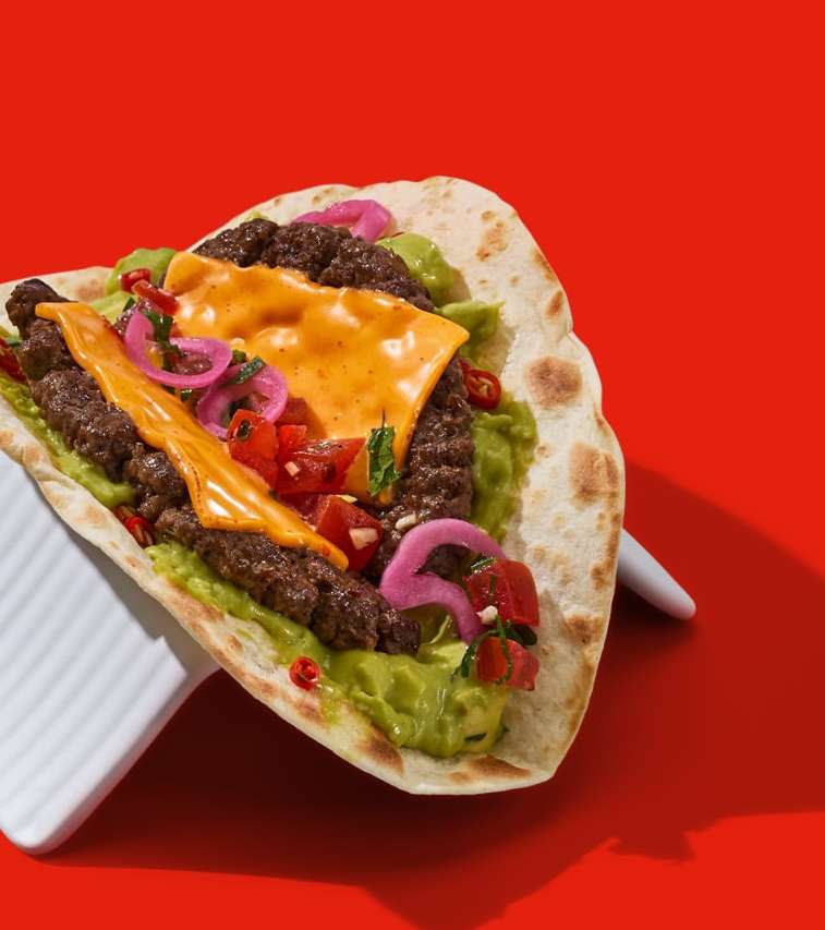 Eye-level close-up of a taco on a white surface featuring seasoned beef, shredded cheddar cheese, guacamole, tomatoes, and pickled onions.