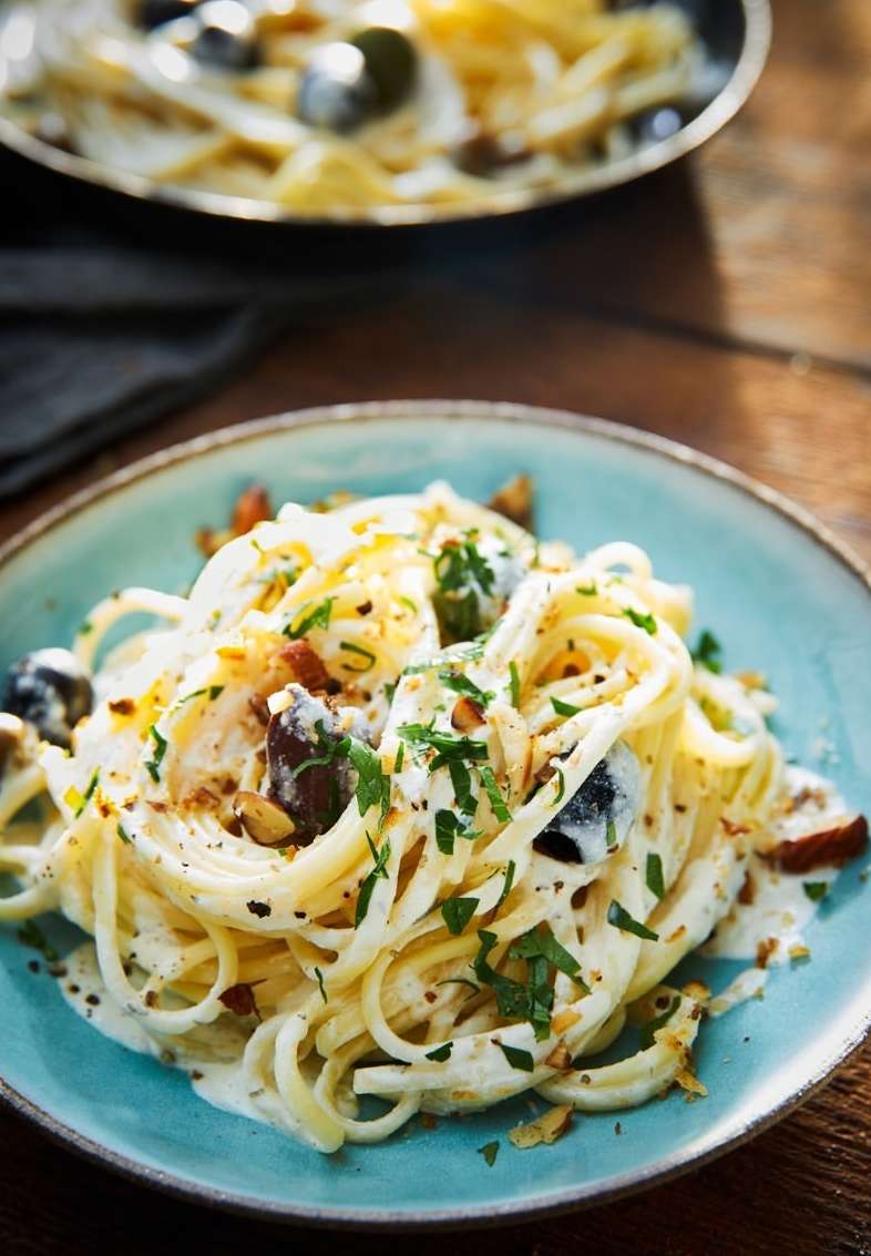 Overhead shot of pasta dish with creamy white sauce, dark olives, and chopped parsley on a light blue plate, set on a wooden surface.