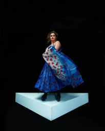 Model in a blue patterned dress standing on a white triangular platform against a black background