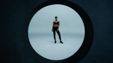 Woman in black athletic top and leggings framed inside a circular opening against a grey background