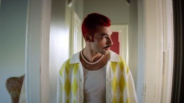 Headshot of a young adult male with a red hairstyle wearing a yellow patterned cardigan and pearl necklace in an indoor setting.