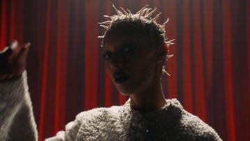 Dramatic close-up portrait of a person with spiky hair and dark lipstick against a red curtain backdrop, lit with a spotlight.