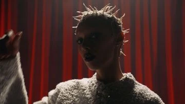 Dramatic close-up portrait of a person with spiky hair and dark lipstick against a red curtain backdrop, lit with a spotlight.