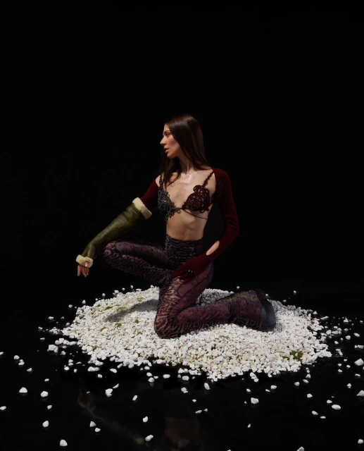 A woman poses in maroon and green patterned bra top and pants, seated on white gravel against a black backdrop in a studio shot.