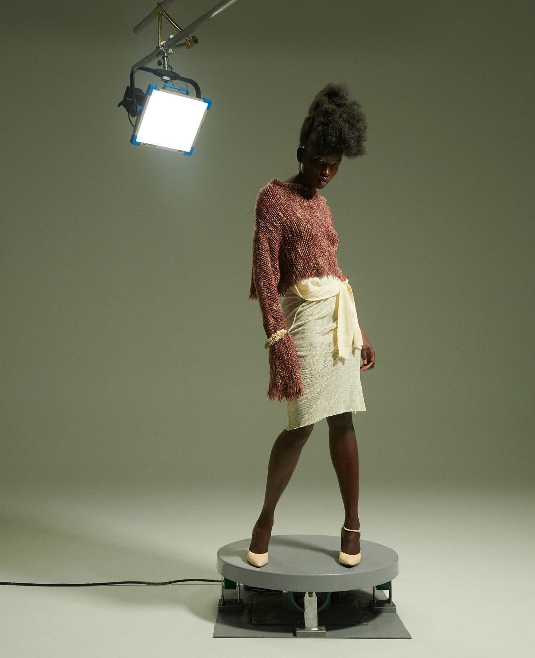 Fashion model poses on rotating platform in a photo studio wearing a sweater and skirt under bright light, showcasing fashion and style.