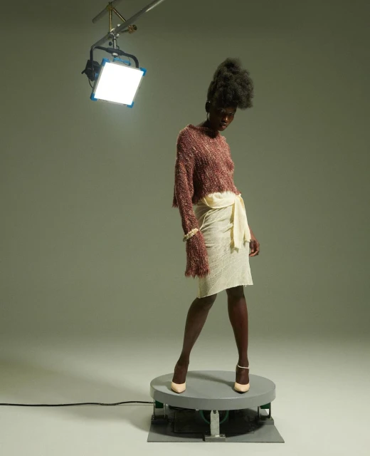 Fashion model poses on rotating platform in a photo studio wearing a sweater and skirt under bright light, showcasing fashion and style.