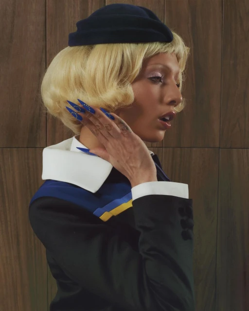 Adult woman in a dark jacket with a white collar and blue hat, posing in front of a brown wooden wall, fashionably dressed.