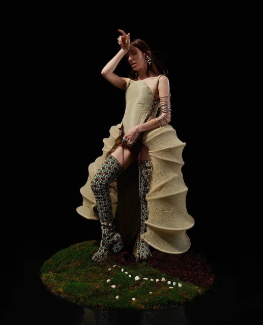 A woman in a gold dress with high patterned boots stands on a mossy platform against a dark backdrop in a fashion or editorial shot.