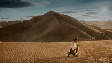 Adult female sits in desert landscape against dunes. Wearing light colored jacket and shorts. Sunny, natural light in outdoor environment.