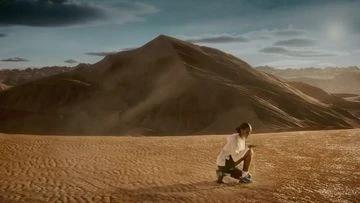 Adult female sits in desert landscape against dunes. Wearing light colored jacket and shorts. Sunny, natural light in outdoor environment.