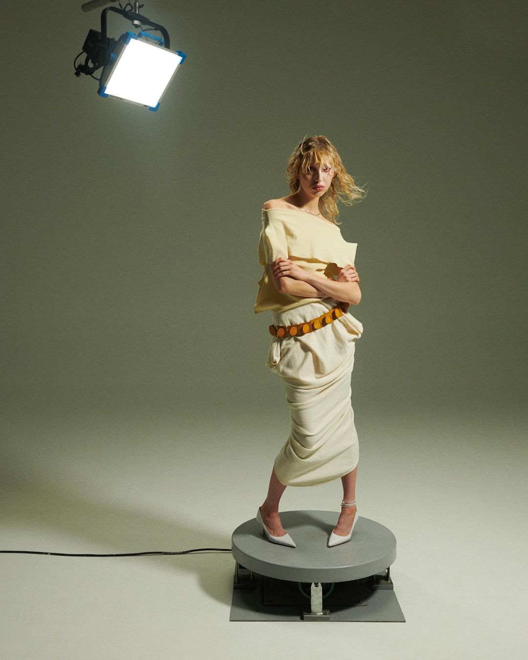 A stylish blonde woman in an off-shoulder top and draped skirt stands on a platform in a studio illuminated by bright lighting.