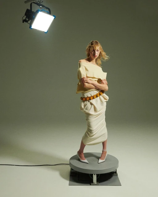 A stylish blonde woman in an off-shoulder top and draped skirt stands on a platform in a studio illuminated by bright lighting.