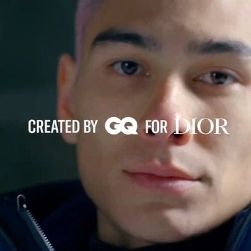 Close-up of a man with short pink hair, wearing a black hooded jacket, set against a dark background, with 'Created by GQ for Dior' text.