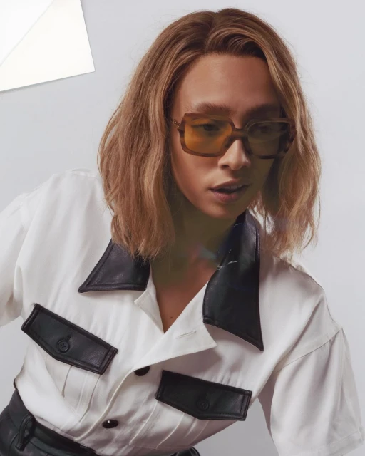 Adult posing in white shirt with black details and amber sunglasses, in a studio environment with soft lighting.