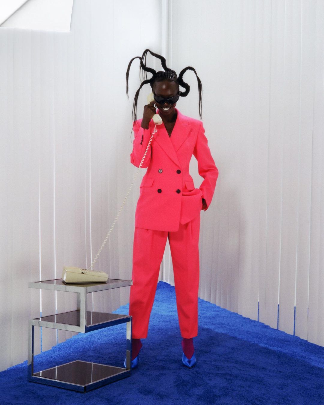 A Black woman in a pink suit poses in front of a white backdrop, holding a beige telephone in a fashion-style editorial photo.