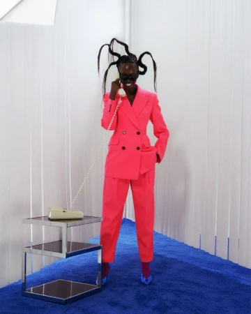 A Black woman in a pink suit poses in front of a white backdrop, holding a beige telephone in a fashion-style editorial photo.