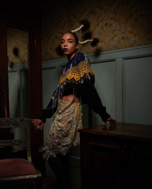 A woman with unique hair poses indoors wearing a cropped sweater with attached chains, and a tan fringed skirt in moody lighting.