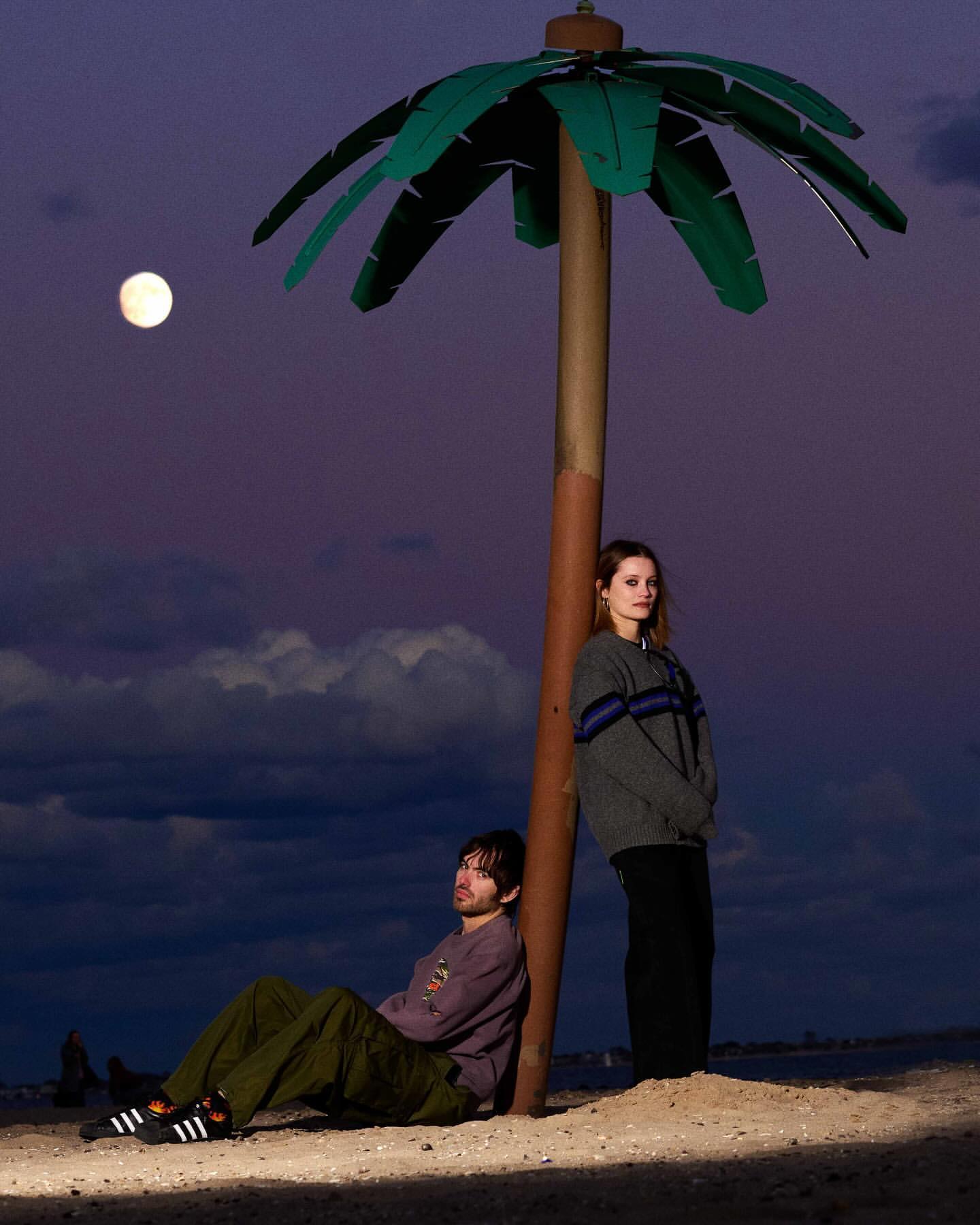 An adult male and female pose on a sandy surface under a faux palm tree at dusk against a sky with a bright moon.