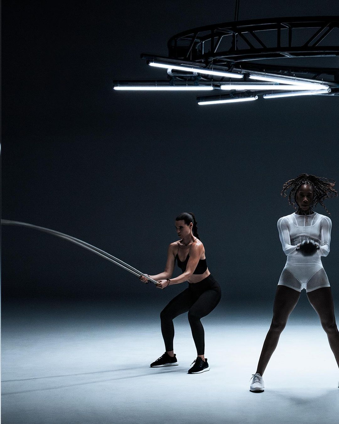 Two women work out in a dark studio with weights and battle ropes, illuminated by a modern geometric lighting fixture above.