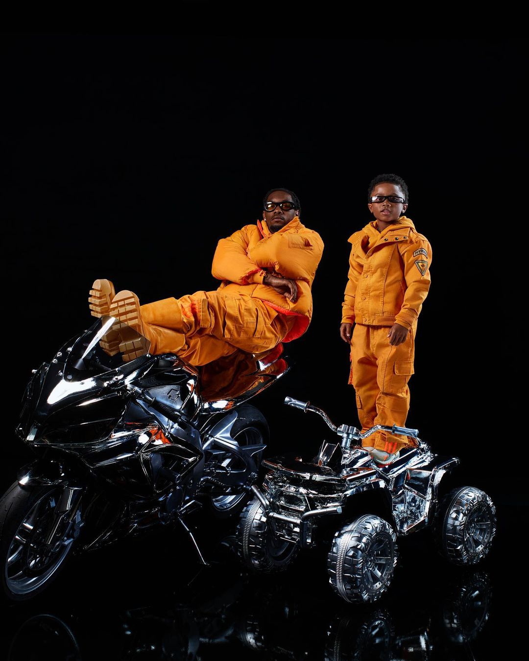 An adult male and a child pose in orange jumpsuits next to shiny motorcycles against a black background in a studio environment.