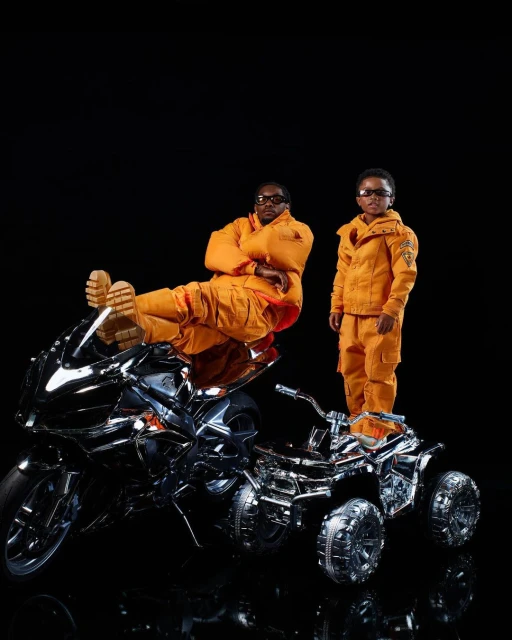 An adult male and a child pose in orange jumpsuits next to shiny motorcycles against a black background in a studio environment.