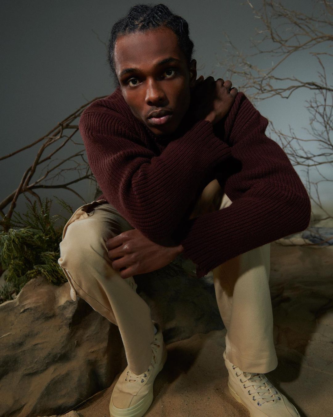 Stylish young man with dark hair, wearing a burgundy sweater and khaki pants, poses outdoors among greenery and rock formations.