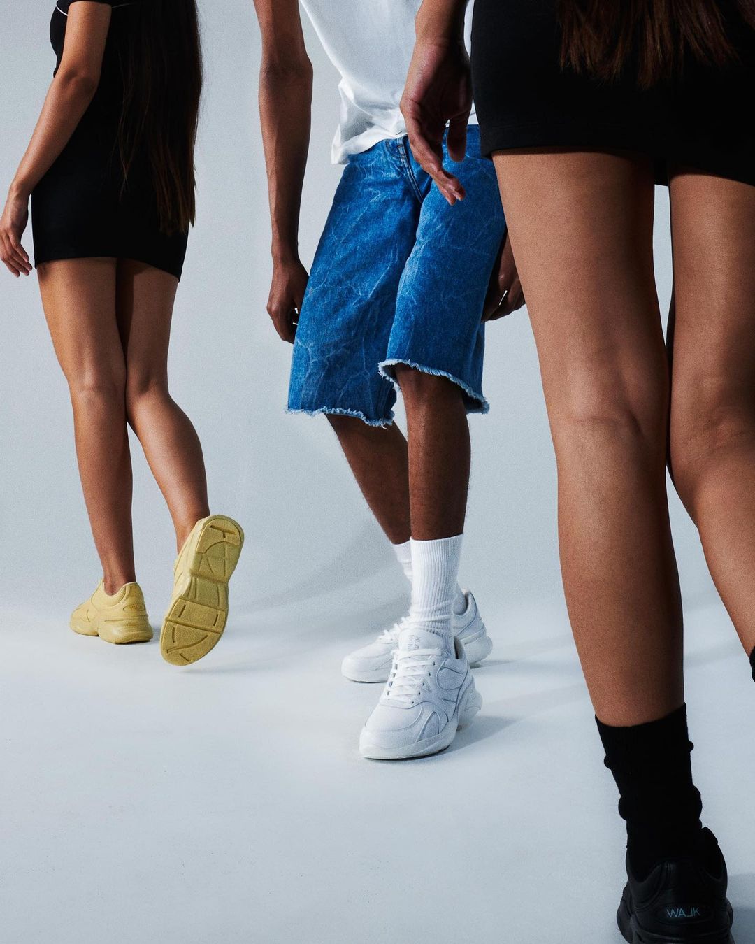 Three fashion models showcase denim shorts and sneakers in a studio setting. The focus is on the shoes and lower body.