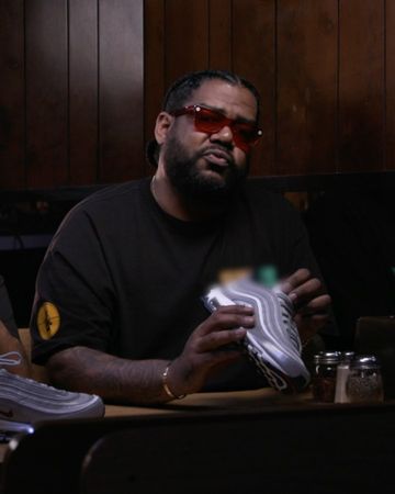 Adult male wearing sunglasses, black tee-shirt holding a grey running shoe, in a casual indoor setting with wood panelling.