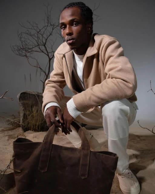 Black male model in beige jacket and white pants squats with brown tote bag in studio setting with rock and branches. Fashion editorial.