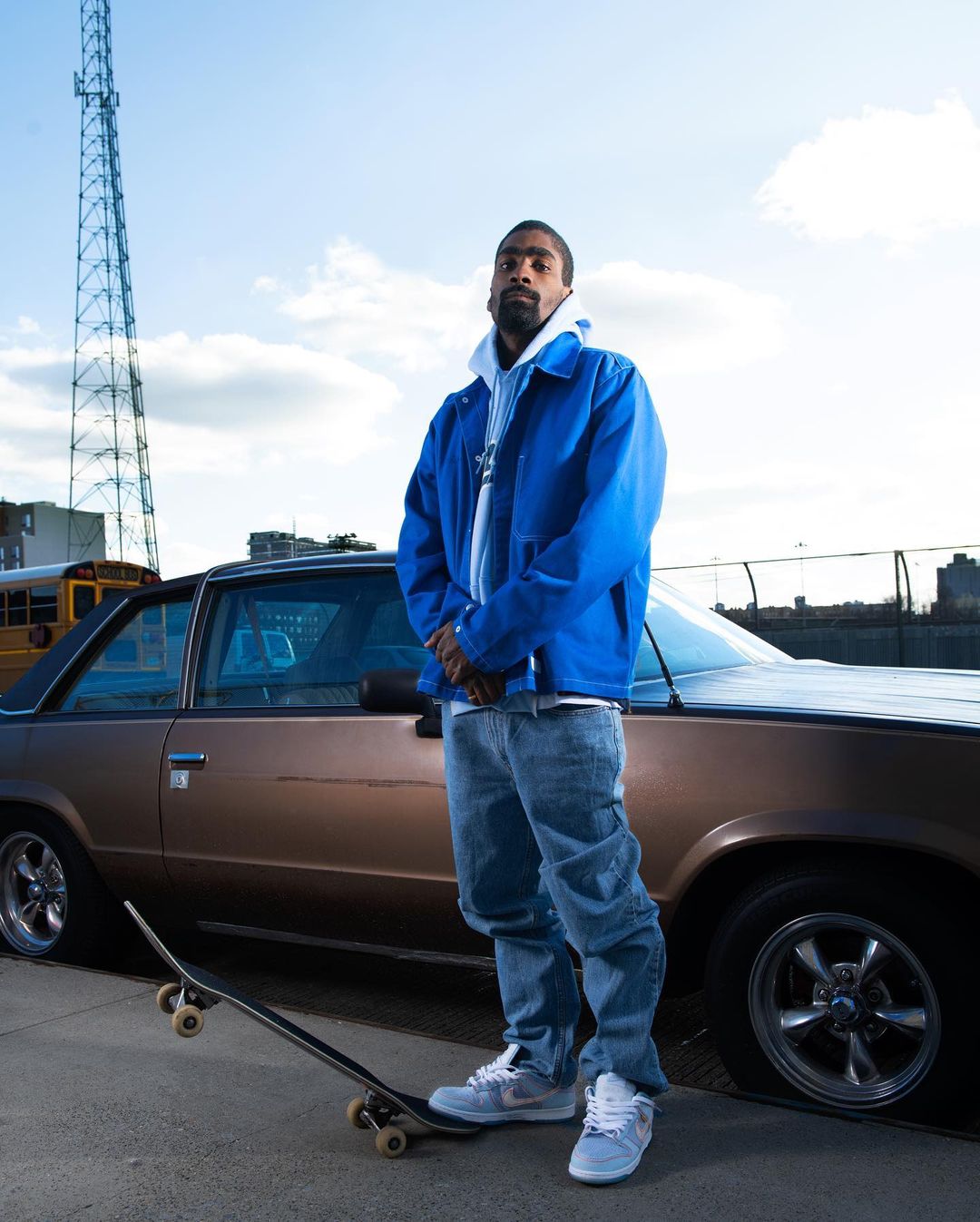 Adult man in blue jacket poses next to vintage car with a skateboard in an urban setting under a blue sky with communication tower.