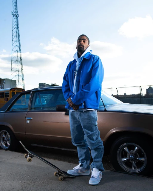 Adult man in blue jacket poses next to vintage car with a skateboard in an urban setting under a blue sky with communication tower.