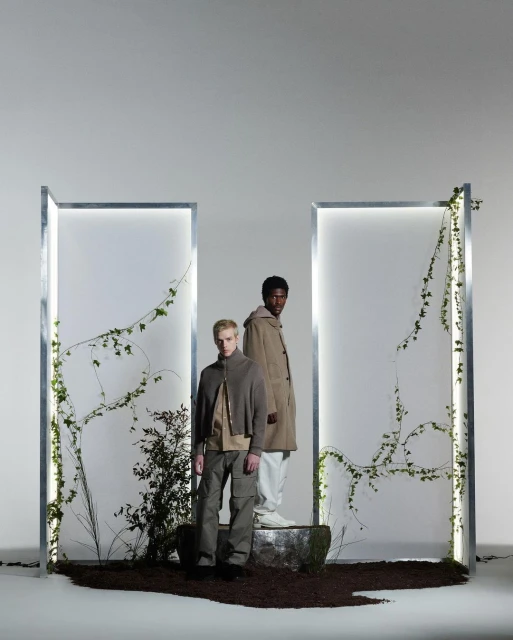 Two fashion models pose in coordinated neutral-toned jackets and pants framed by vertical light boxes with climbing ivy in studio.