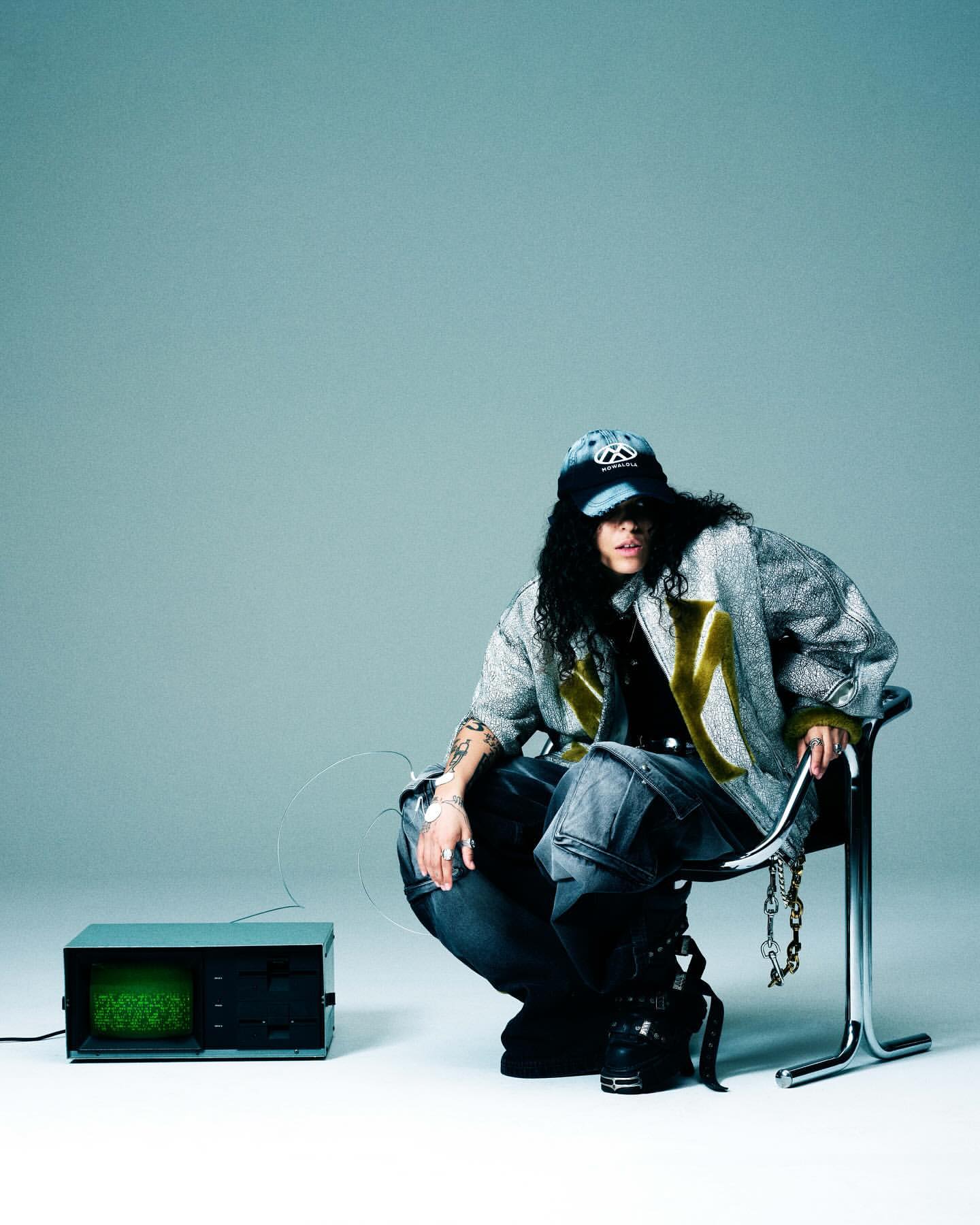 Person crouches near an old television wearing jeans, boots, baseball cap, and silver jacket in a studio with a grey background.