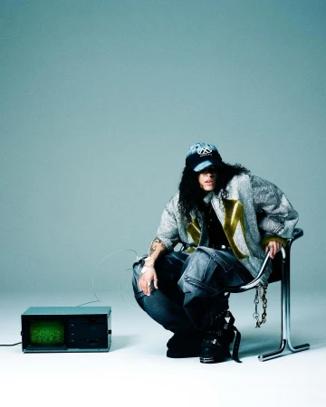 Person crouches near an old television wearing jeans, boots, baseball cap, and silver jacket in a studio with a grey background.