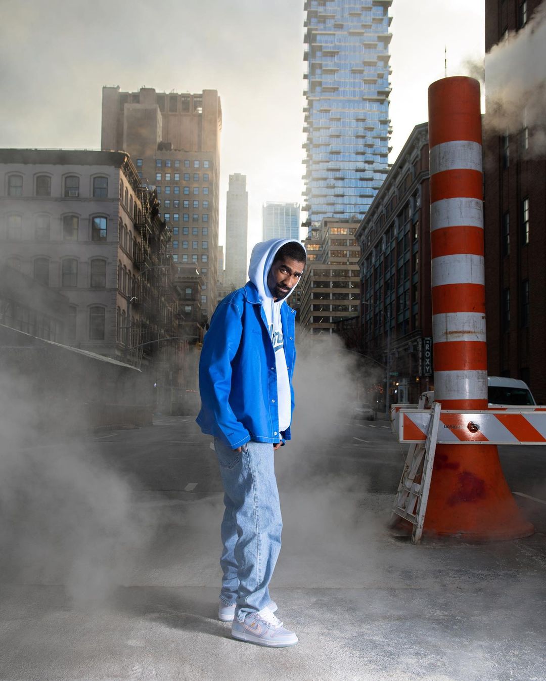 A man in a blue jacket and jeans stands in a foggy urban setting with tall buildings and an orange and white striped safety barrier.