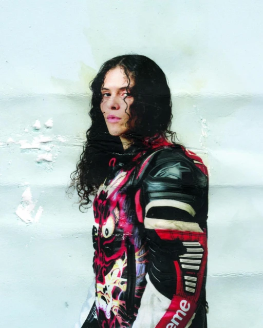 Woman with long, dark curly hair poses in a black and red racing suit against a weathered, light blue wall with paint remnants.