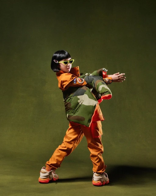 Young child strikes a pose wearing a camouflage jacket, orange pants and yellow glasses against an olive green backdrop in studio shot.