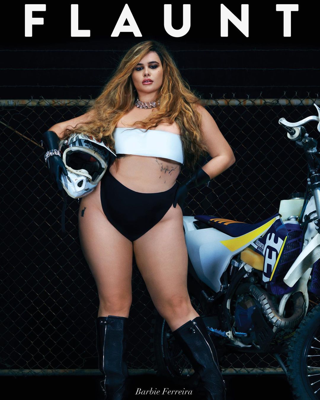 A woman in minimal clothing poses with a dirt bike next to a chain-link fence under bright lighting for a fashion magazine cover shoot.