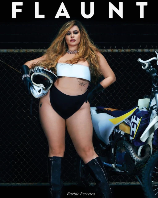 A woman in minimal clothing poses with a dirt bike next to a chain-link fence under bright lighting for a fashion magazine cover shoot.
