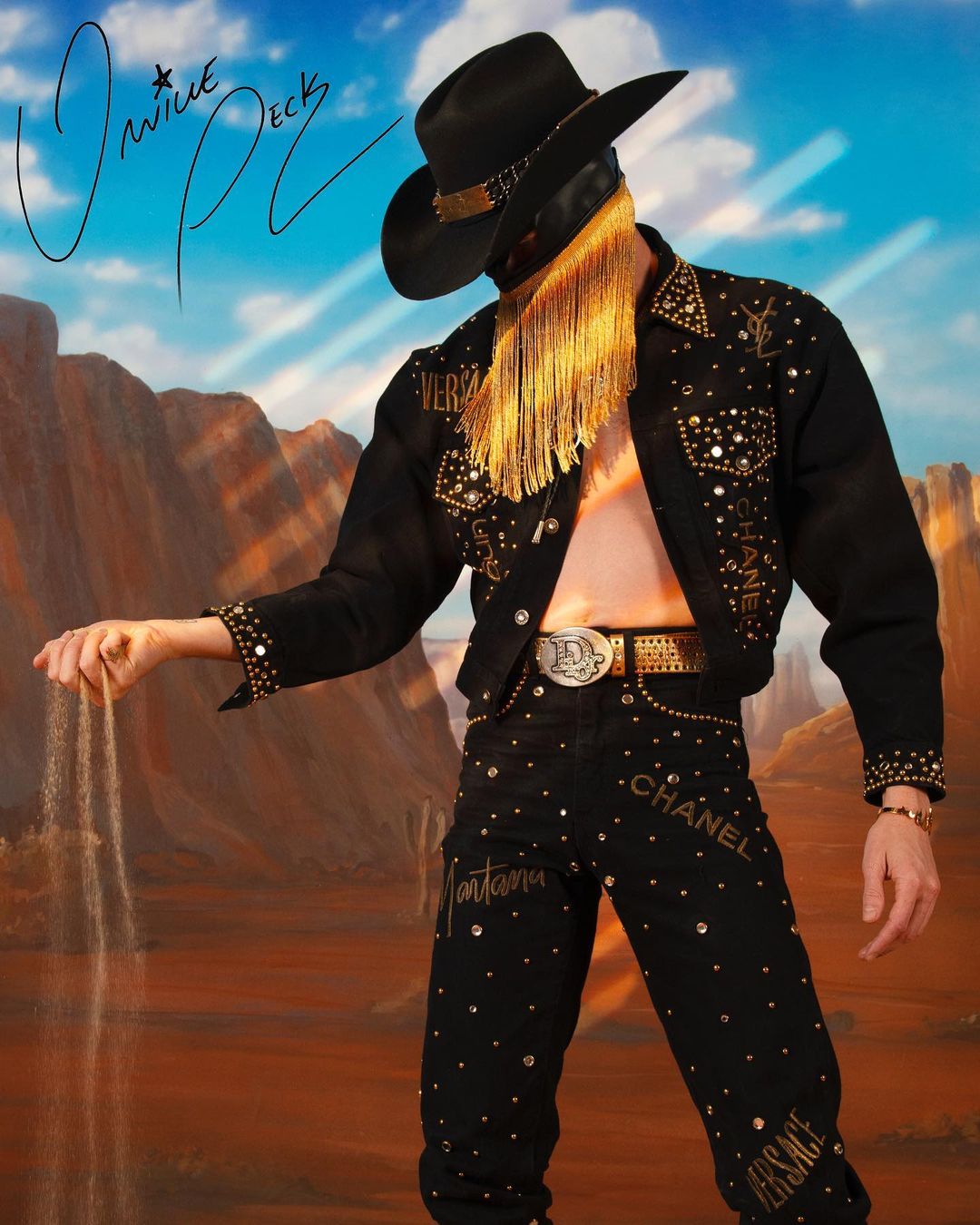 Orville Peck poses in a black cowboy hat, mask with gold fringe, and custom rhinestone-studded black jacket and pants on a desert landscape.