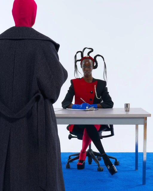 Woman with creative hairstyle seated at desk with pen and paper, another person in a coat stands nearby in a brightly lit room.