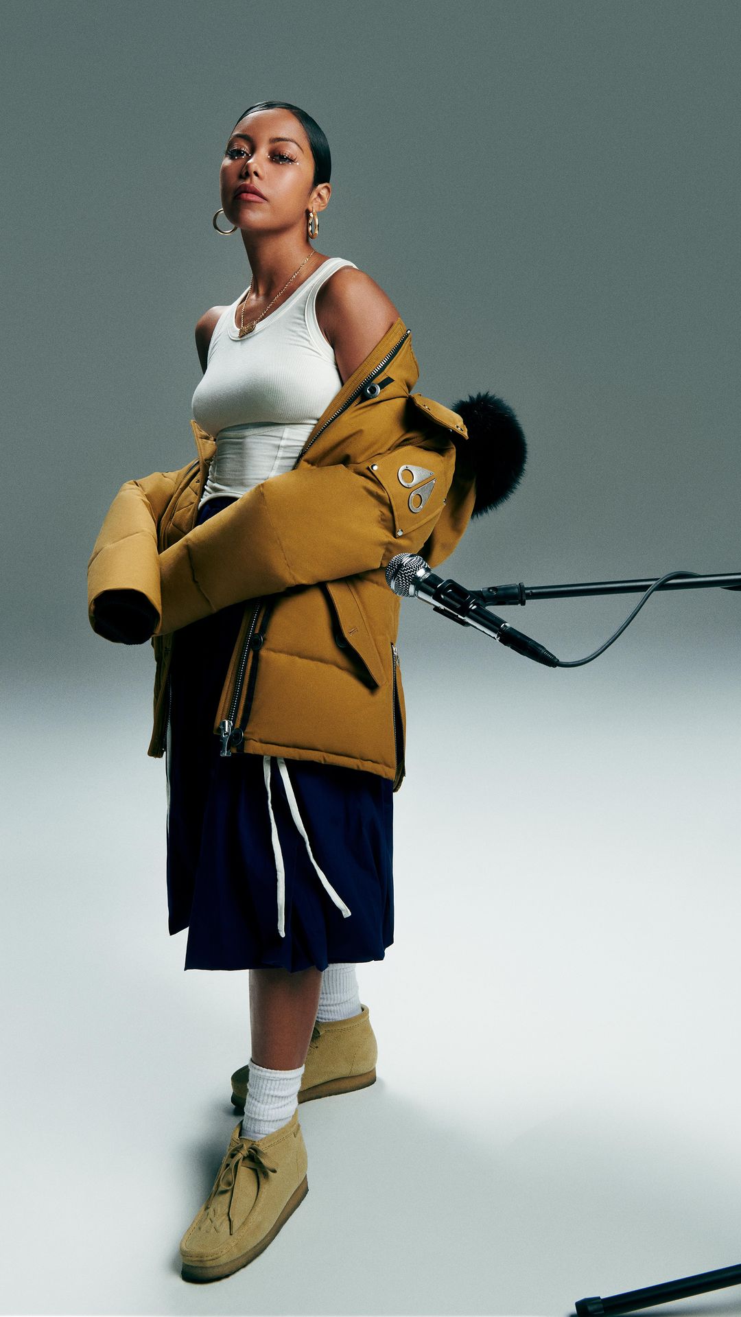 A fashionable woman stands in a studio wearing a tank top, navy skirt, tan boots, and a mustard-colored puffer jacket draped around her.